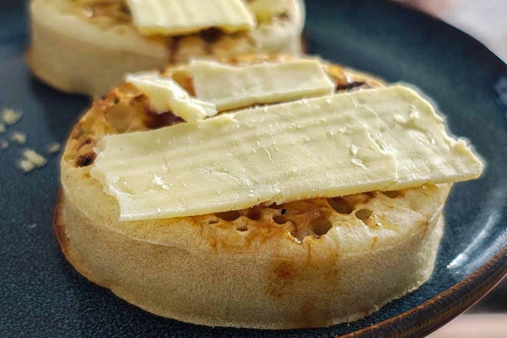 Paahdettu crumpet, jonka päällä on Marmite-levitettä ja viipaleita vaaleaa juustoa. Taustalla näkyy toinen samanlainen crumpet lautasella.