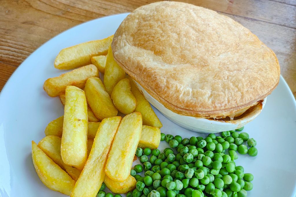 Perinteinen brittiläinen chicken pie tarjoiltuna ranskalaisten perunoiden ja vihreiden herneiden kanssa valkoisella lautasella.
