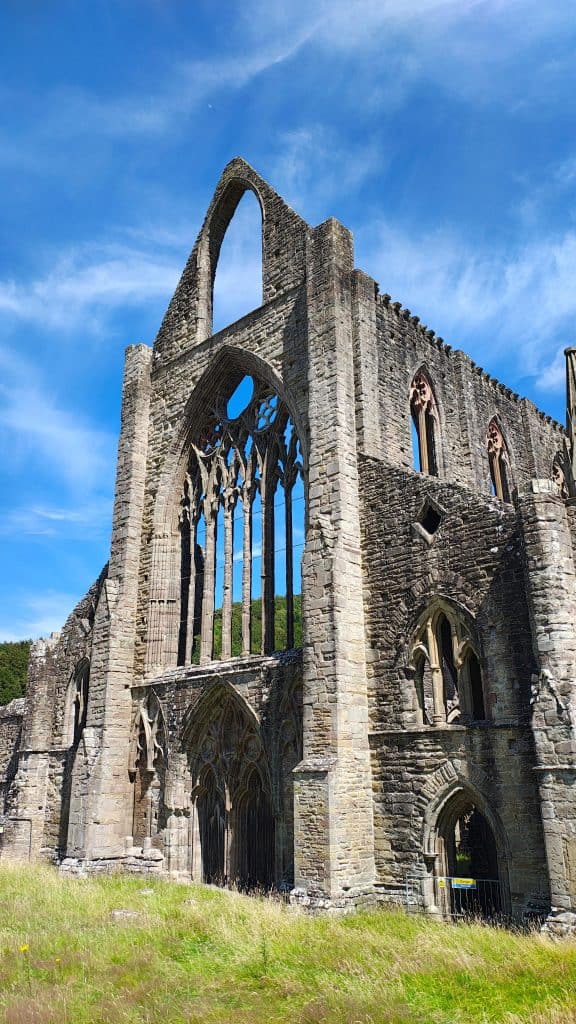 Tintern Abbeyn korkeat goottilaiset rauniot sinistä taivasta vasten. Kivirakennuksessa on suuria kaari-ikkunoita ja koristeellisia ikkuna-aukkoja.